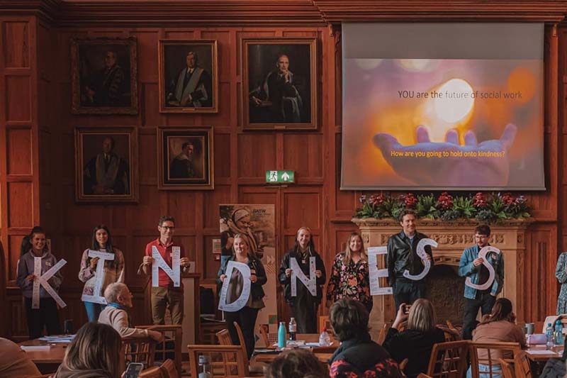 students standing in a row against a wood panelled wall, holding large letters making up the word kindness